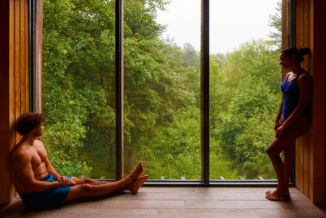 Man and a woman looking out at the forest from inside the Treetop Sauna.